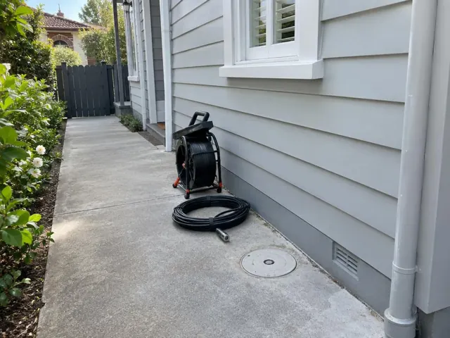 Capped round inspection cover beside a coiled drain camera reel on a concrete side path by weatherboard house in Cammeray