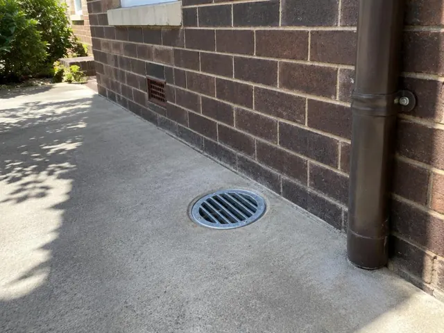 Outdoor concrete path beside brick wall with a round metal drain grate and downpipe in Cremorne