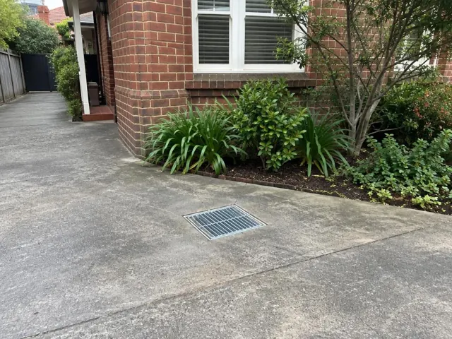 Concrete driveway with a seated stormwater grate beside a brick house and garden in Cremorne